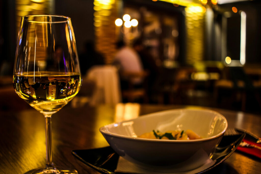 Photo of a glass of white wine next to a plate in a darkened dining room.